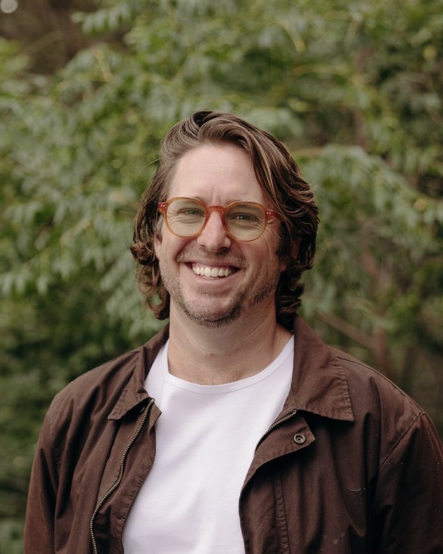 Dan Hamilton smiling in tortoiseshell glasses, a white tee and a brown jacket, with green trees behind him